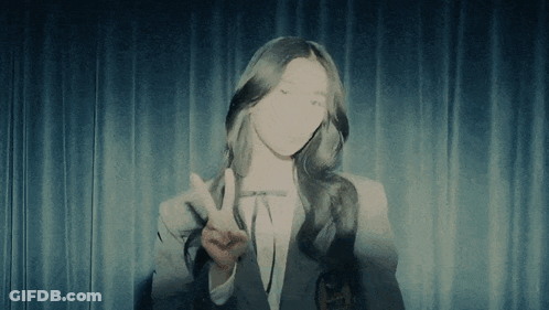 A Woman Giving A Peace Sign In Front Of A Curtain GIF