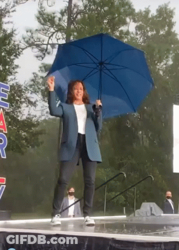 A Woman Holding A Blue Umbrella Stands On A Stage In Front Of A Sign That Says " Har " GIF