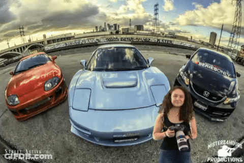 A Woman Holding A Camera Stands In Front Of A Series Only Car And A Red Car GIF