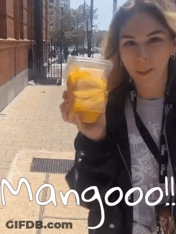 A Woman Holding A Cup Of Mango Juice With The Words Mangooo Written On The Sidewalk GIF