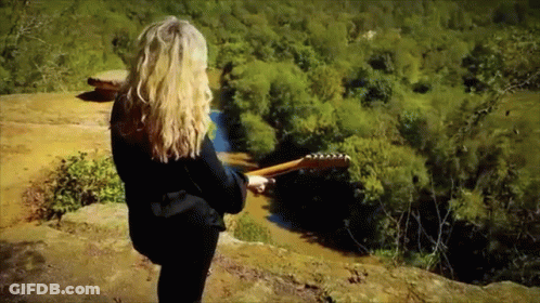 A Woman Holding A Guitar Standing On A Cliff Overlooking A River GIF