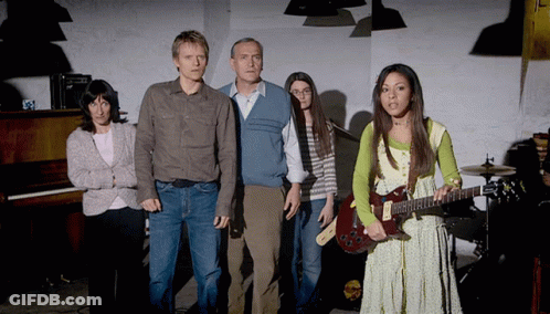 A Woman Holding A Guitar Stands In Front Of A Group Of People GIF