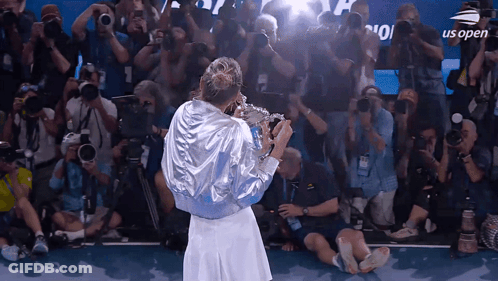 A Woman Holding A Trophy In Front Of A Crowd With Us Open On The Screen GIF