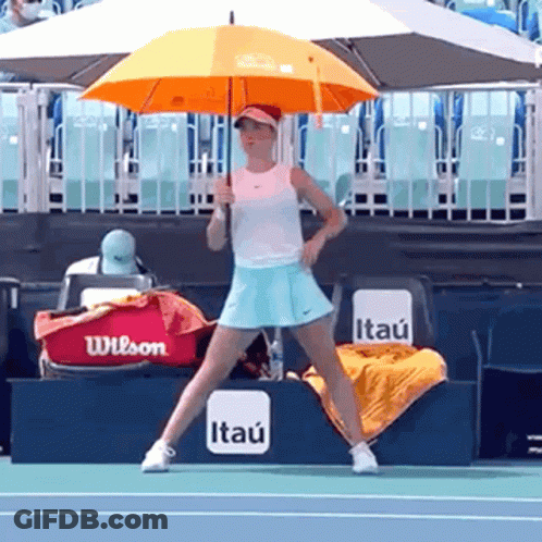 A Woman Holding An Orange Umbrella On A Tennis Court With A Wilson Bag In The Background GIF
