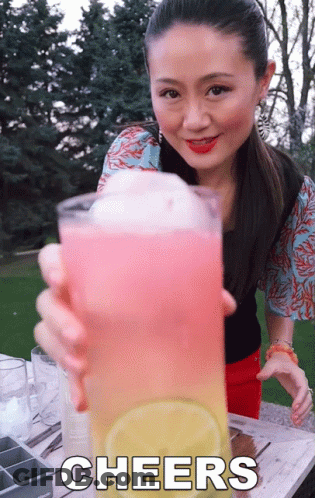 A Woman Holds Up A Glass Of Pink And Yellow Liquid With The Words Cheers Written Below Her GIF