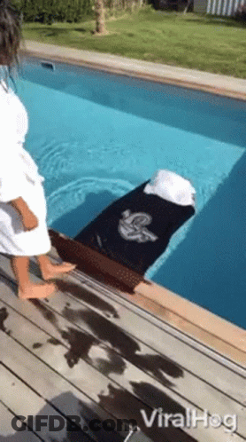 A Woman In A Bathrobe Is Standing Next To A Swimming Pool With A Coffin In It . GIF