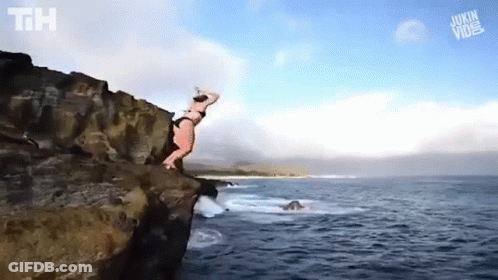 A Woman In A Bikini Is Jumping Off Of A Cliff Into The Ocean . GIF