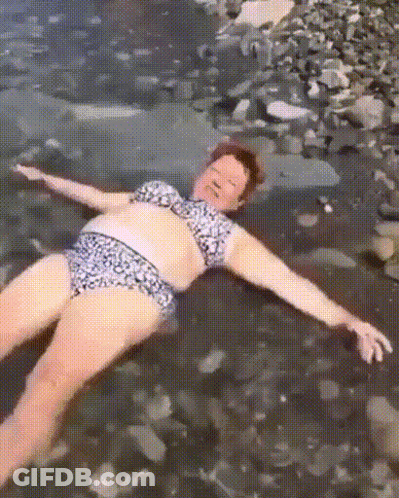 A Woman In A Bikini Is Laying On A Rocky Beach With Her Arms Outstretched . GIF