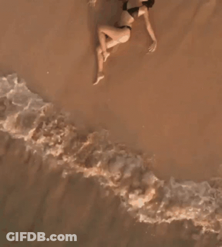 A Woman In A Bikini Is Laying On The Beach Near The Water GIF