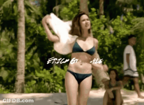 A Woman In A Bikini Is Standing On A Beach Holding A Towel And The Words " Free By Me " Are Visible GIF