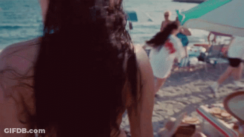 A Woman In A Bikini Is Standing On A Beach Looking At The Ocean GIF