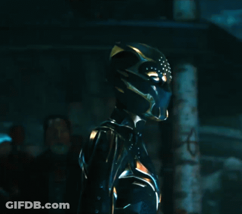 A Woman In A Black And Gold Costume With A Helmet On Stands In A Dark Room GIF