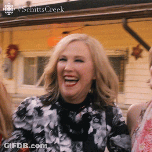 A Woman In A Black And White Floral Dress Is Laughing In Front Of A Sign That Says Schitts Creek GIF