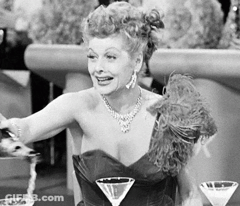 A Woman In A Black And White Photo Is Sitting At A Table With Two Martini Glasses . GIF