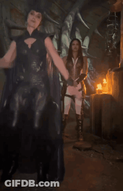A Woman In A Black Cape Stands Next To A Woman In A White Dress GIF