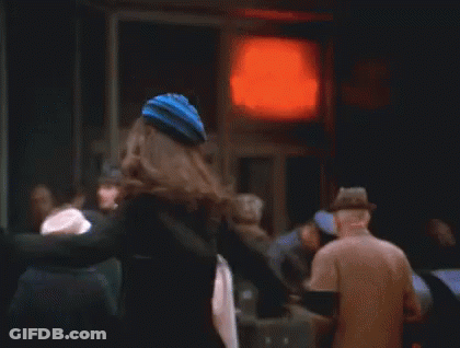 A Woman In A Black Coat And Hat Is Standing In Front Of A Red Light . GIF