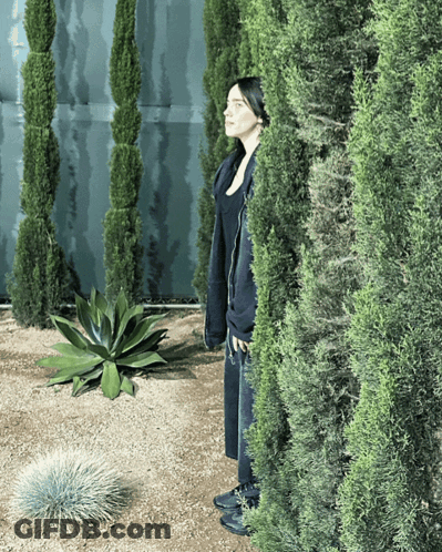 A Woman In A Black Coat Is Standing Behind A Bush GIF