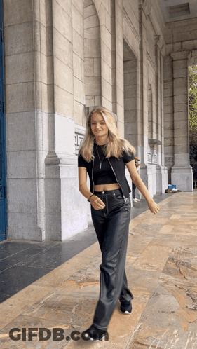 A Woman In A Black Crop Top And Black Leather Pants Walks In Front Of A Building GIF