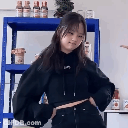 A Woman In A Black Crop Top Is Standing In Front Of A Blue Shelf . GIF