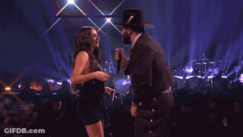 A Woman In A Black Dress Is Dancing On A Stage With A Man In A Cowboy Hat GIF