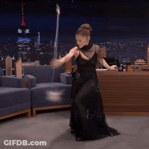 A Woman In A Black Dress Is Dancing With A Wand In Front Of The Empire State Building GIF