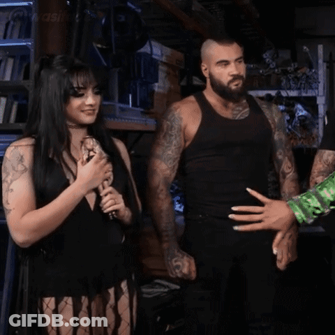 A Woman In A Black Dress Is Holding A Microphone And Smiling While A Man Stands Behind Her . GIF