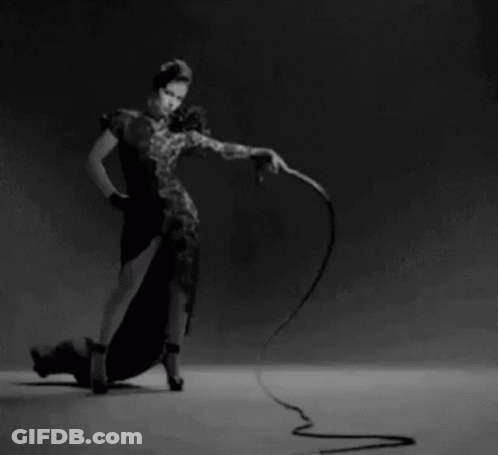 A Woman In A Black Dress Is Holding A Whip In Her Hand GIF