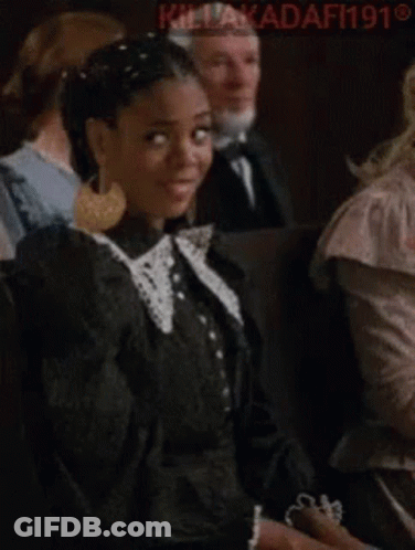A Woman In A Black Dress Is Making A Funny Face While Sitting In A Crowd Of People . GIF