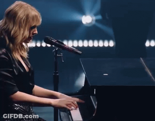 A Woman In A Black Dress Is Playing A Piano And Singing Into A Microphone GIF