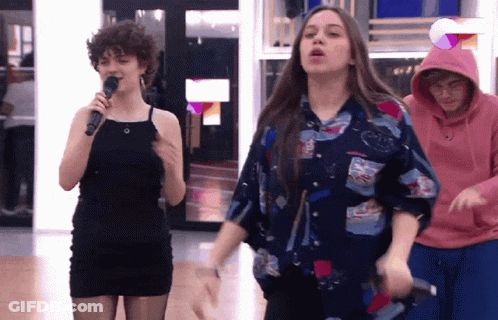 A Woman In A Black Dress Is Singing Into A Microphone While A Man In A Pink Hoodie Watches GIF