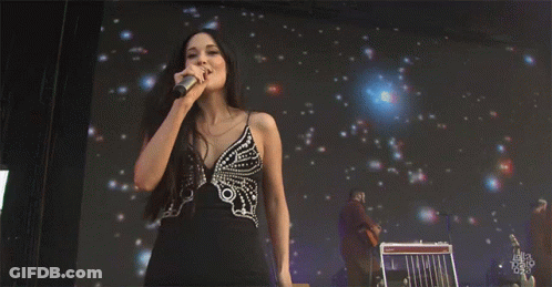 A Woman In A Black Dress Is Singing Into A Microphone While Standing In Front Of A Screen That Says Ella Talk GIF
