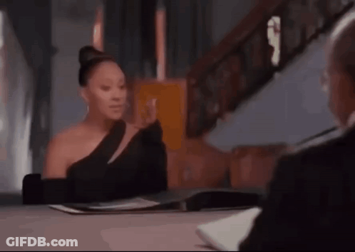 A Woman In A Black Dress Is Sitting At A Table With A Man . GIF