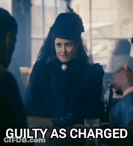 A Woman In A Black Dress Is Sitting At A Table With A Sign That Says Guilty As Charged GIF
