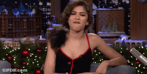 A Woman In A Black Dress Is Sitting In Front Of A Christmas Tree And Making A Funny Face . GIF