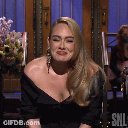 A Woman In A Black Dress Is Sitting In Front Of A Sign That Says Snl On It GIF