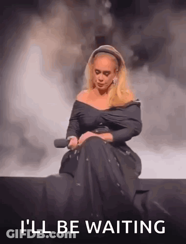 A Woman In A Black Dress Is Sitting On A Stage Holding A Microphone And The Words `` I 'Ll Be Waiting '' GIF