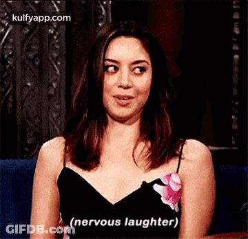 A Woman In A Black Dress Is Smiling And Saying `` Nervous Laughter '' While Sitting On A Couch . GIF