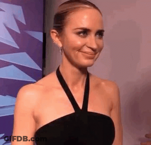 A Woman In A Black Dress Is Smiling In Front Of A Purple Wall . GIF