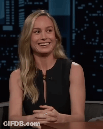 A Woman In A Black Dress Is Smiling With Her Hands Folded In Front Of Her GIF