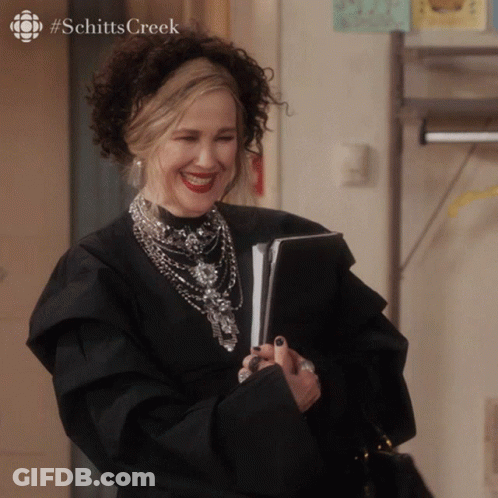 A Woman In A Black Dress Is Smiling With #Schitts Creek On The Bottom Right GIF