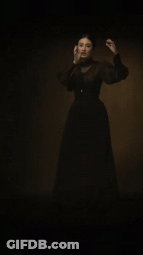 A Woman In A Black Dress Is Standing In A Dark Room With Her Arms Outstretched . GIF