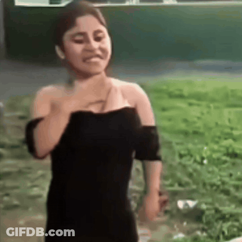 A Woman In A Black Dress Is Standing In A Field With Her Hand On Her Chest . GIF