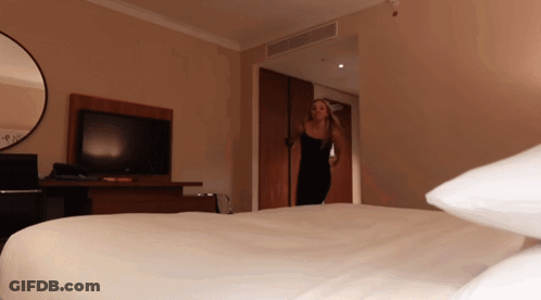 A Woman In A Black Dress Is Standing In A Hotel Room Next To A Bed GIF