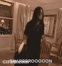 A Woman In A Black Dress Is Standing In A Living Room Holding A Bag And A Purse . GIF