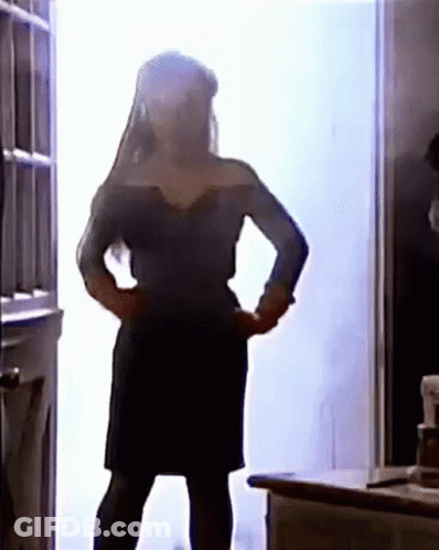 A Woman In A Black Dress Is Standing In Front Of A Door With Her Hands On Her Hips . GIF