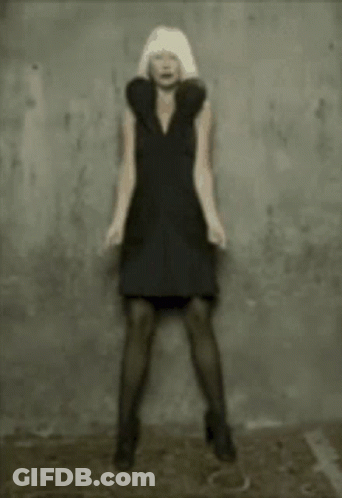 A Woman In A Black Dress Is Standing In Front Of A Gray Wall GIF