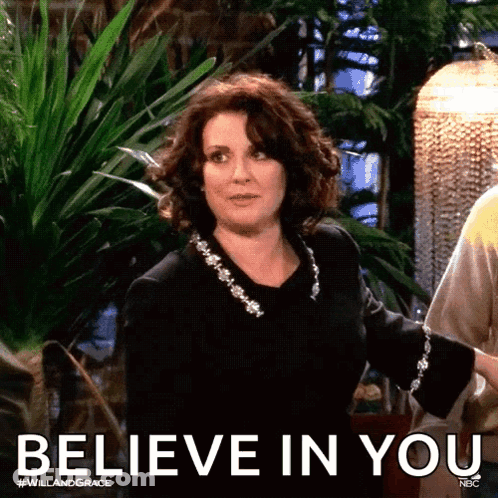 A Woman In A Black Dress Is Standing In Front Of A Sign That Says Believe In You GIF