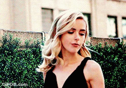 A Woman In A Black Dress Is Standing Next To A Hedge . GIF