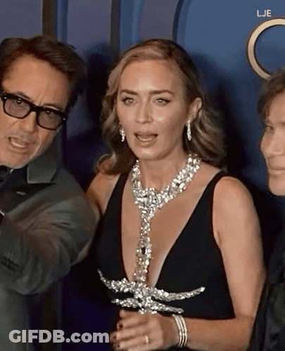 A Woman In A Black Dress Is Standing Next To A Man In Glasses GIF