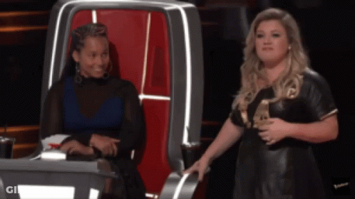 A Woman In A Black Dress Is Standing Next To Another Woman In A Chair GIF
